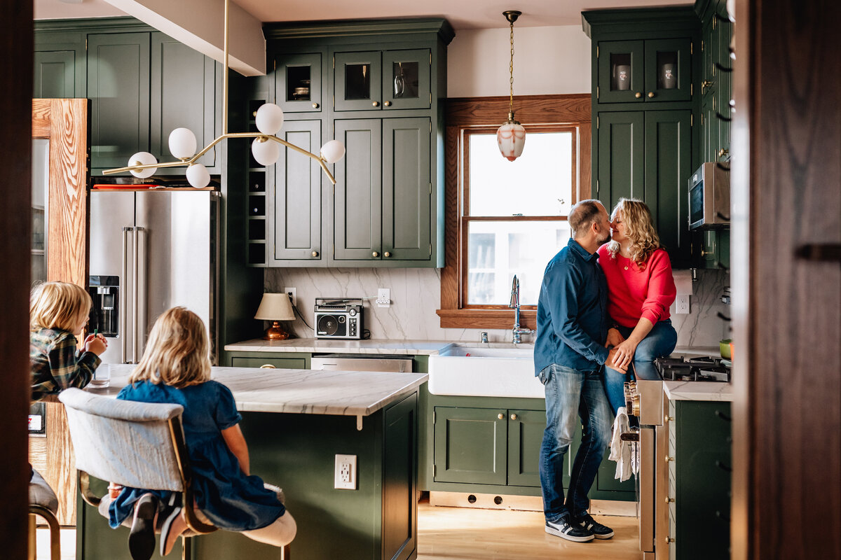 A warm kitchen scene with a couple sharing a tender moment near the stove, while two children sit at the kitchen island, watching with quiet curiosity.