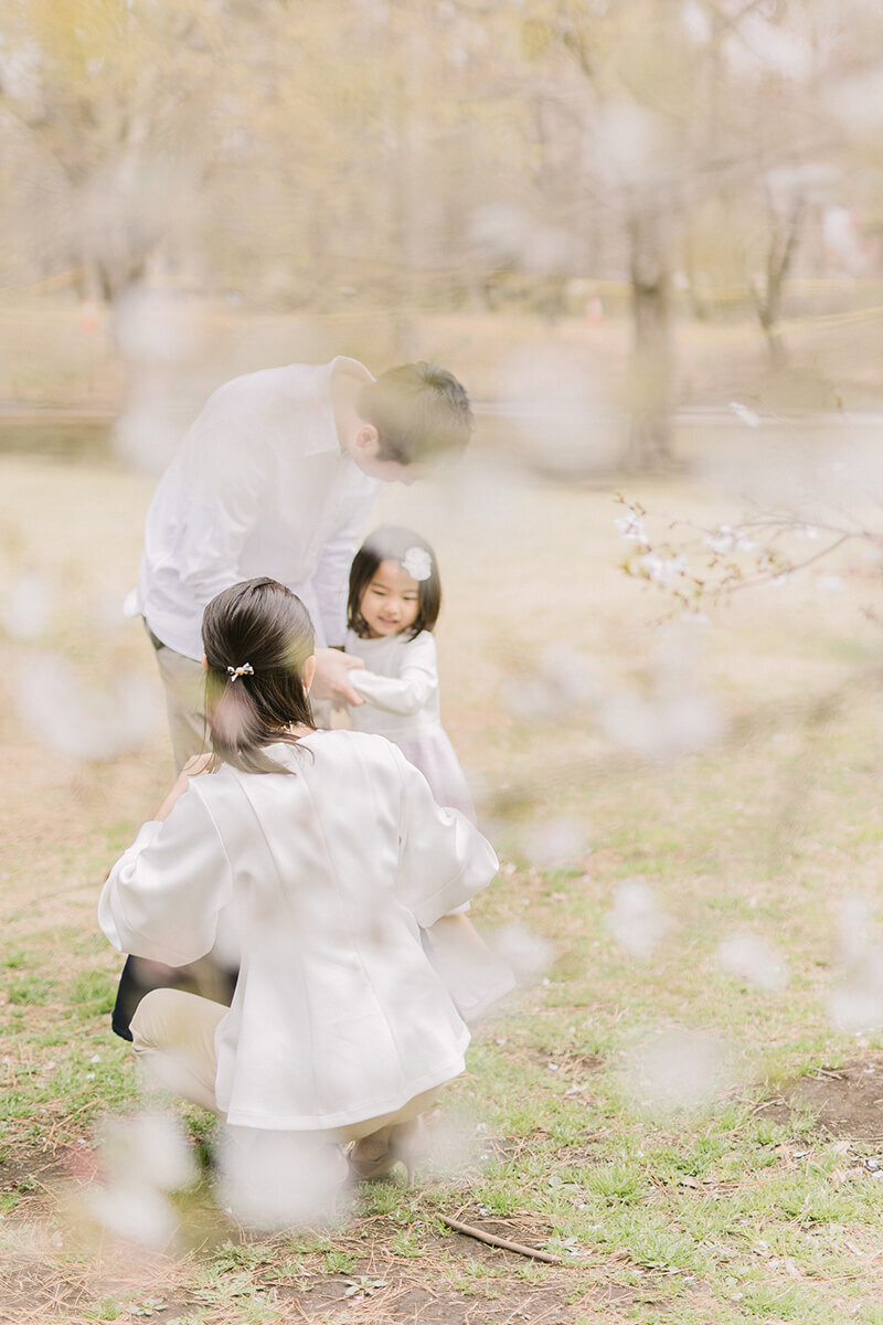 桜の木越しに、女の子に寄り添うママとパパが優しい雰囲気のオシャレな家族写真。相模原市の公園で撮影。