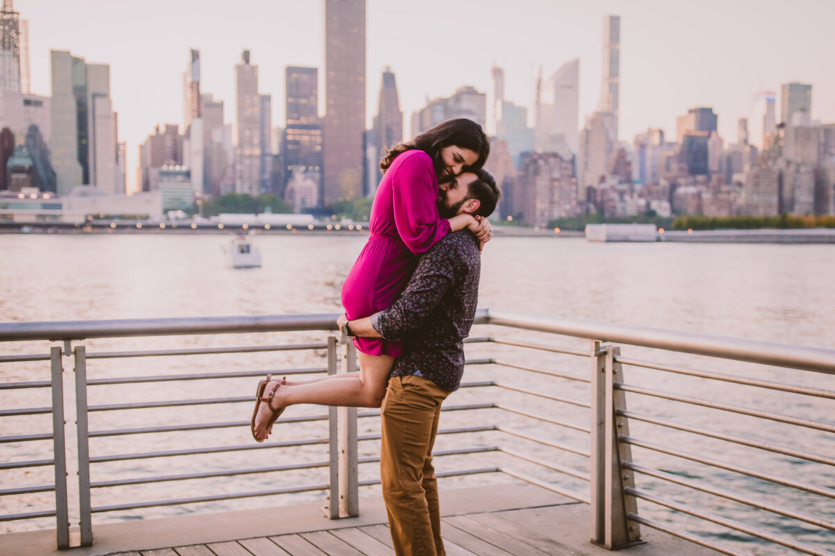 Groom picks up bride during their engagement session at the promenade at gantry plaza in nyc with the skyline in the back