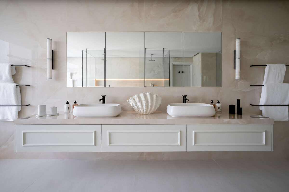 Large double-sink in a primary bathroom, with a focus on light, neutral tones. The sinks are white on a light marble counter, with a large mirror above them.