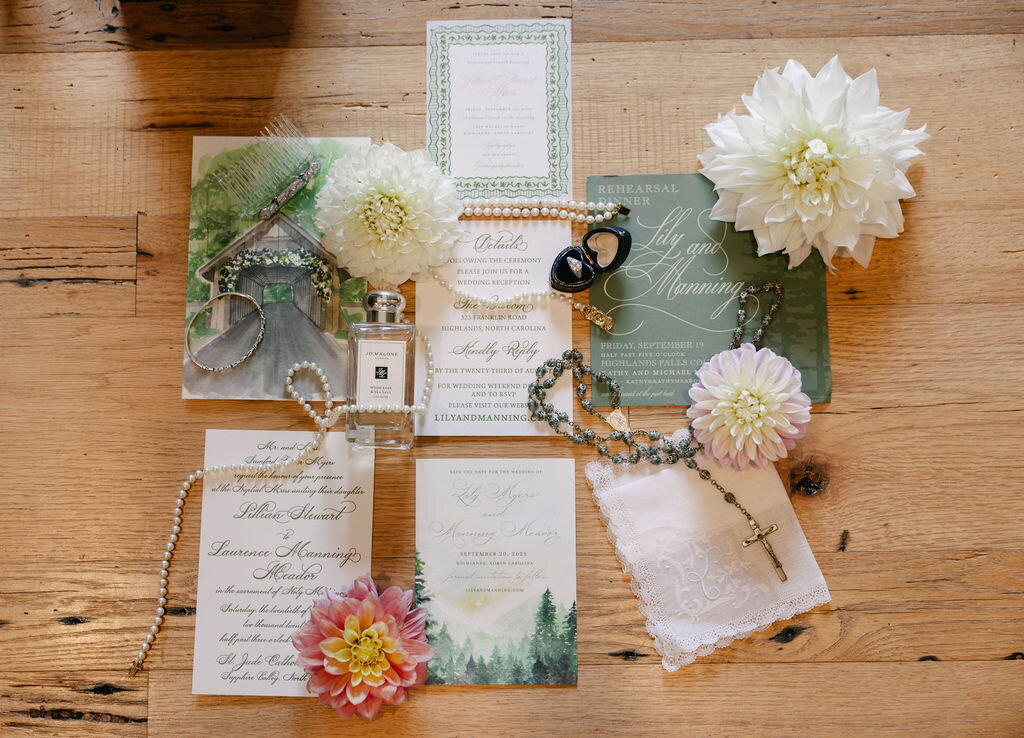 Wedding invitation suite and detail flatlay with florals, jewelry, and rosary for a Highlands, NC wedding at The Bascom.