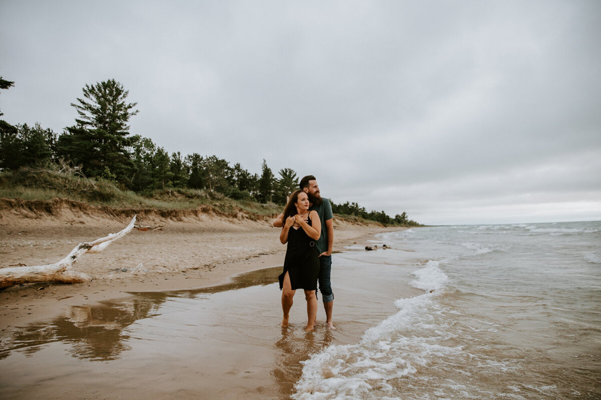 Man and woman standing on Grand Bend beach for engagement photoshoot. The woman's back is to the mans, he is holding her with his arm over her shoulder. Both are looking off in the distance.