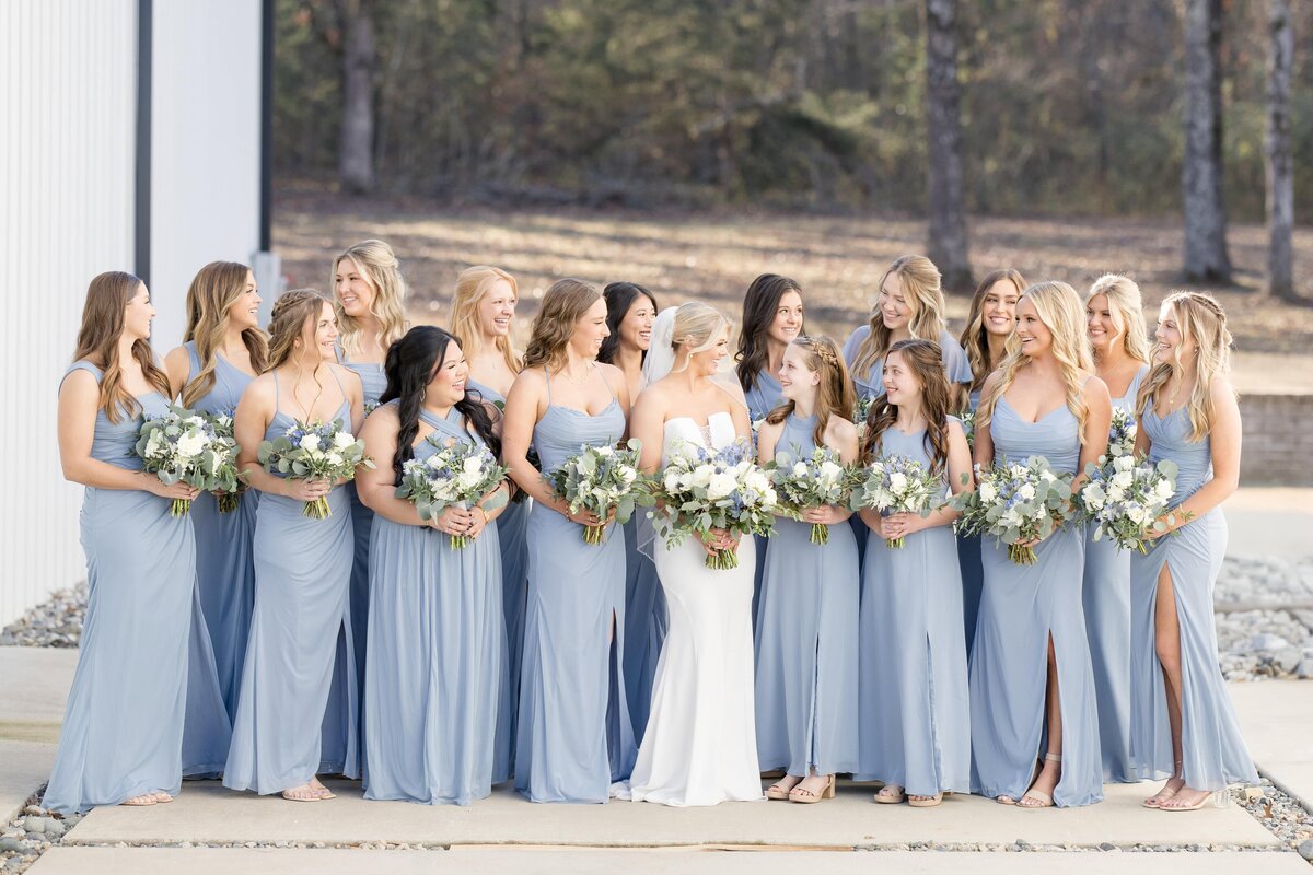 A large wedding party in blue dresses stand holding their bouquets looking at the bride captured by an Arkansas wedding photographer