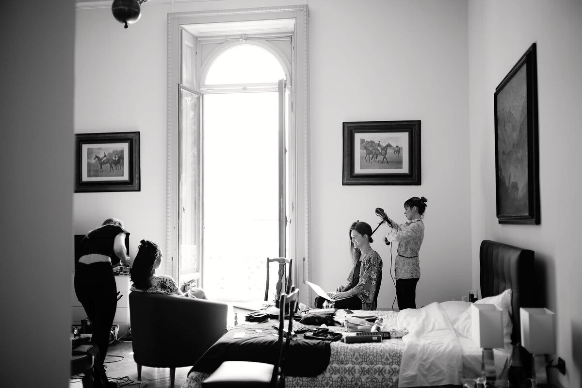 Four women inside a huge room with an oversized window, two of them having their hair and make-up done by the other two