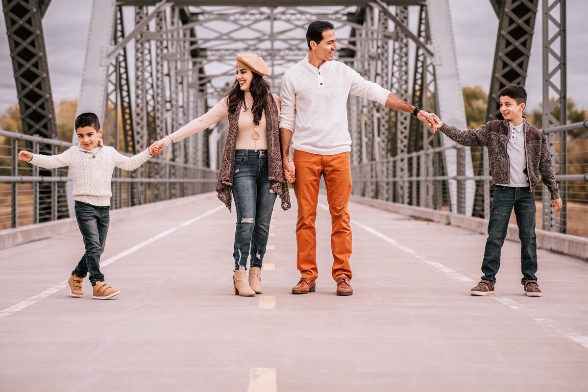 family of four holding hands and walking on a steel bridge. The parents are in the middle, with their two young sons on either side, all wearing coordinated fall outfits.