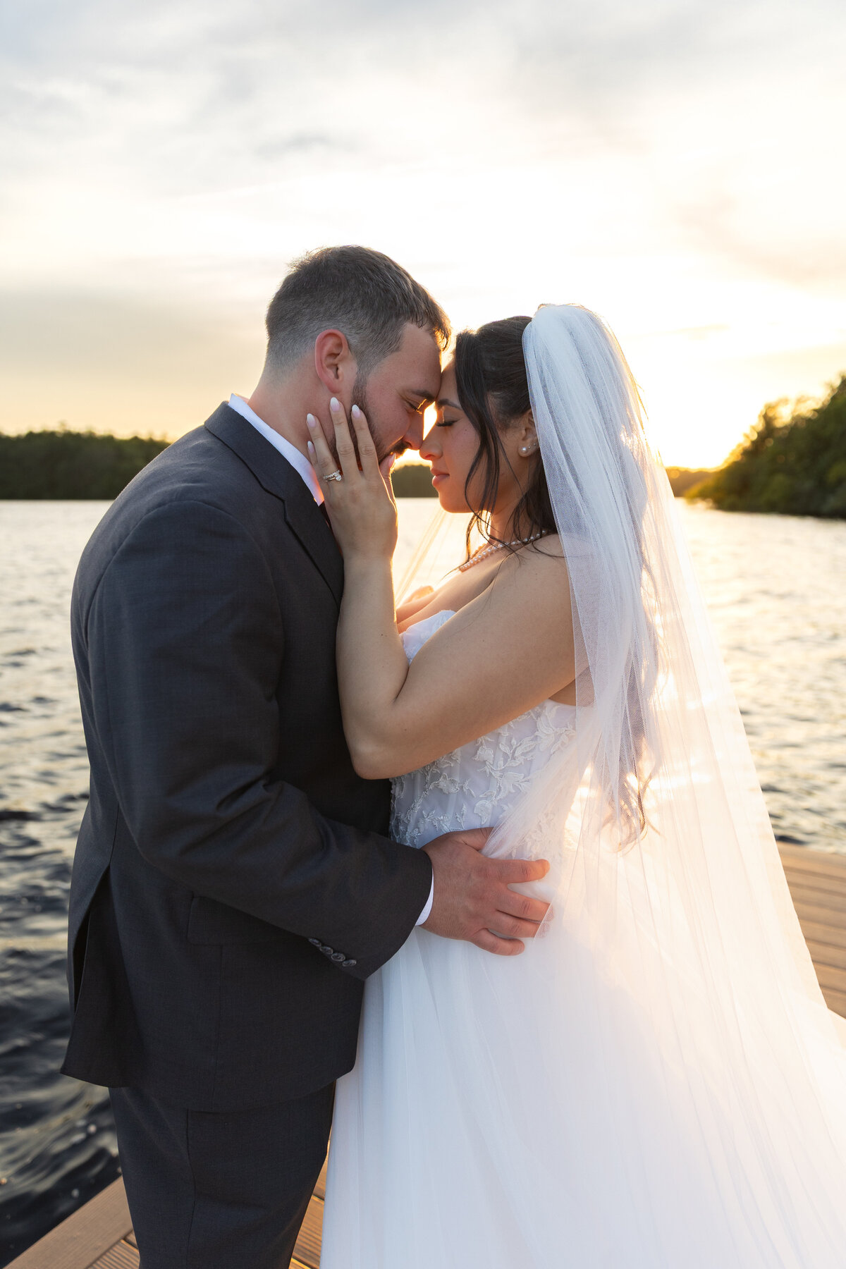 The Lakehouse Inn MA  | Kelsey Sheehan Photography Timeless Rhode Island Weddings | A bride and groom embrace tenderly on a lakeside dock at sunset. She wears a veil and white dress; he is in a dark suit. The scene exudes warmth and romance.
