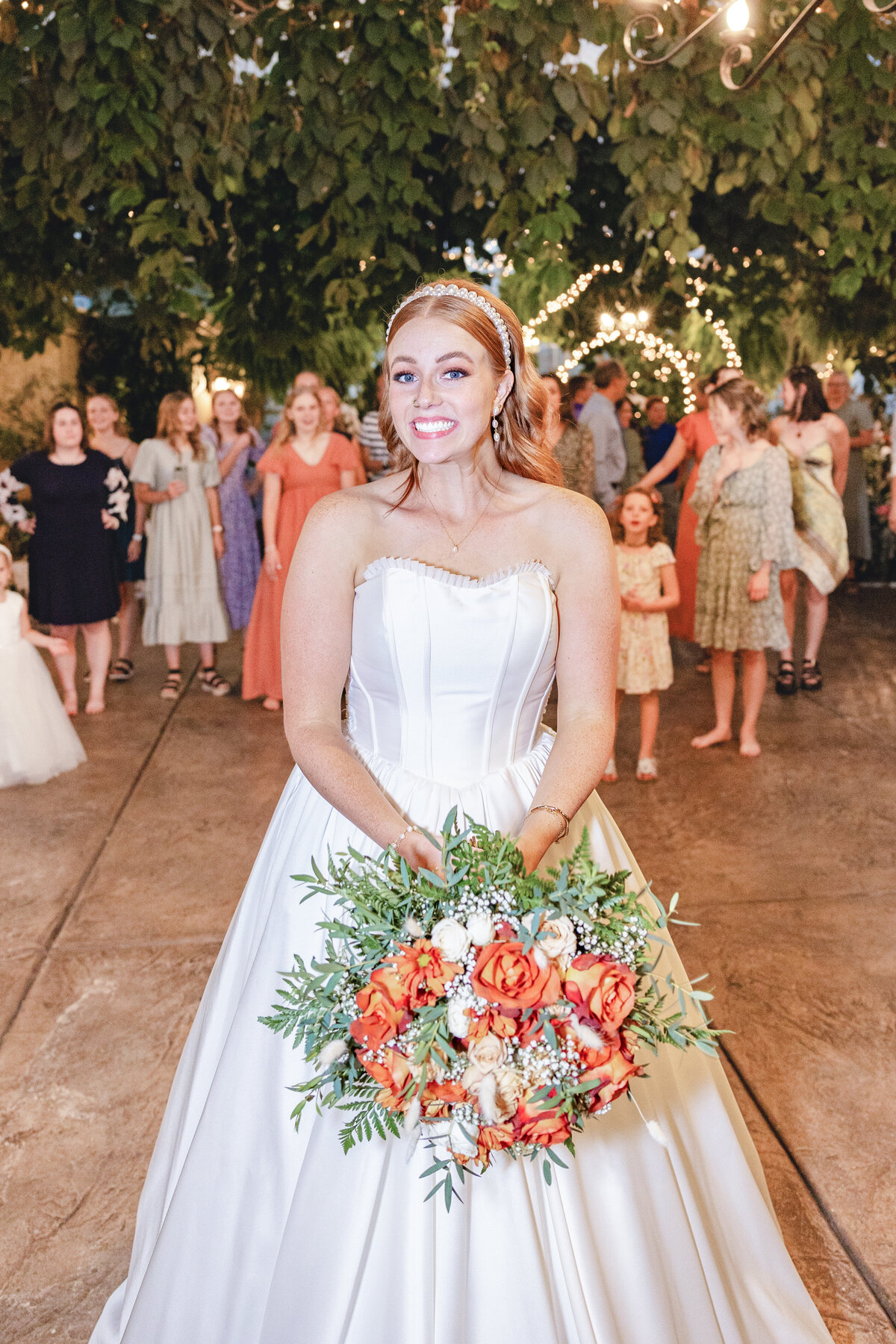 The bride, with a beaming smile, holds her bouquet of orange and white flowers, preparing to toss it to a group of eager single guests.
