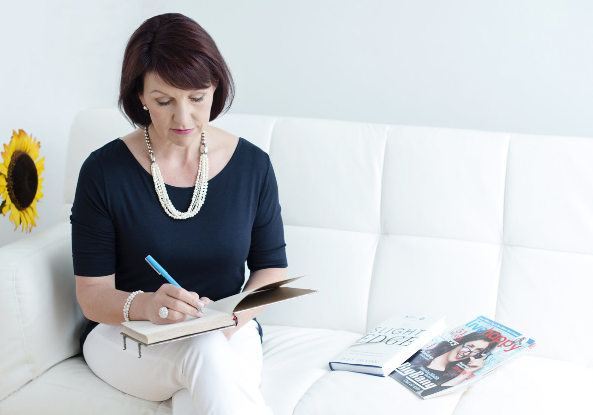business portrait of  leader woman  sitting on desk  and  writing making notes  with magazine on the sofa  wearing white pants and  navy top shot in oakville