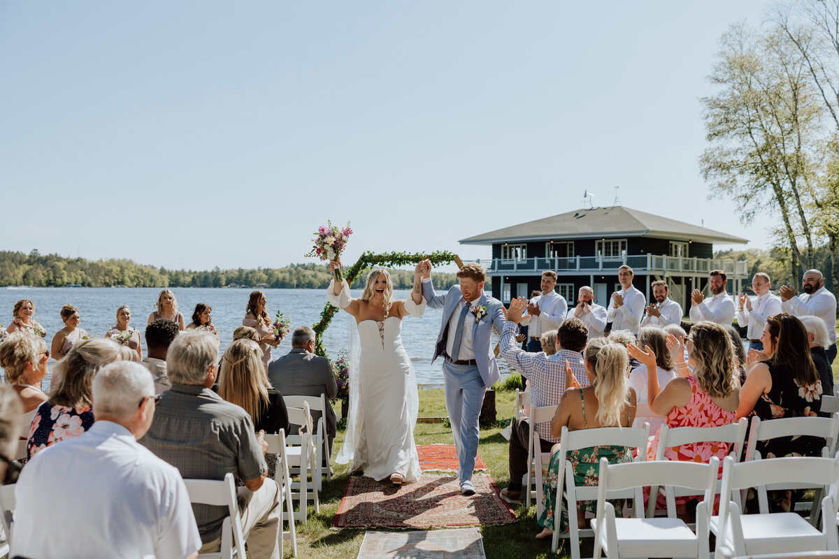 Bride & groom cheering holding hands walking down the aisle after getting married outdoors by a lake
