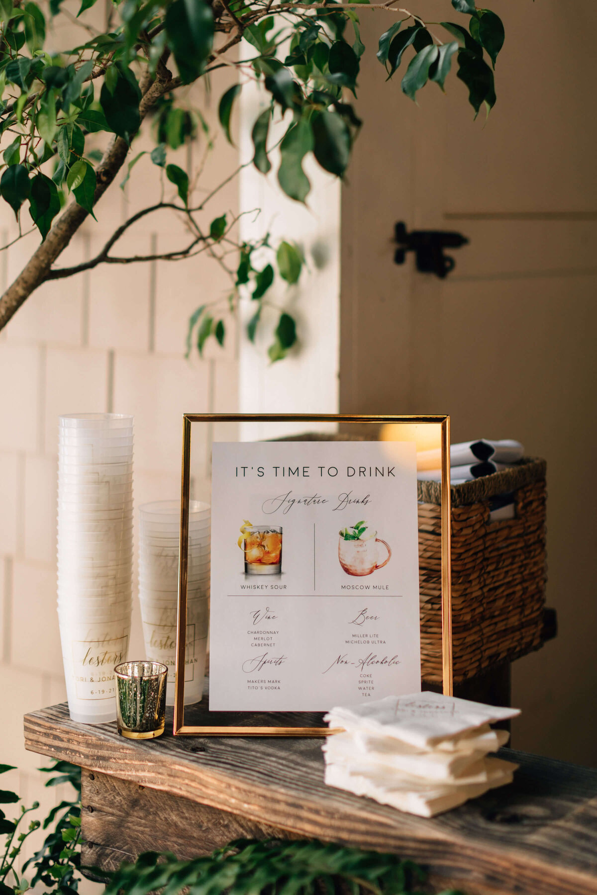 A framed drink menu sits on a wooden bar top beside stacked plastic cups, napkins, and a wicker basket, all outdoors next to a leafy plant and light-colored wall.