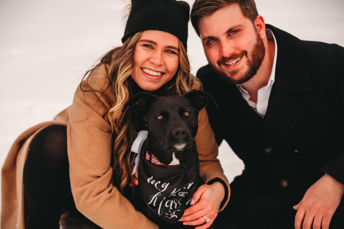 A smiling engaged couple poses with their black dog wearing a wedding bandana during a cozy winter engagement session in Erie, PA.