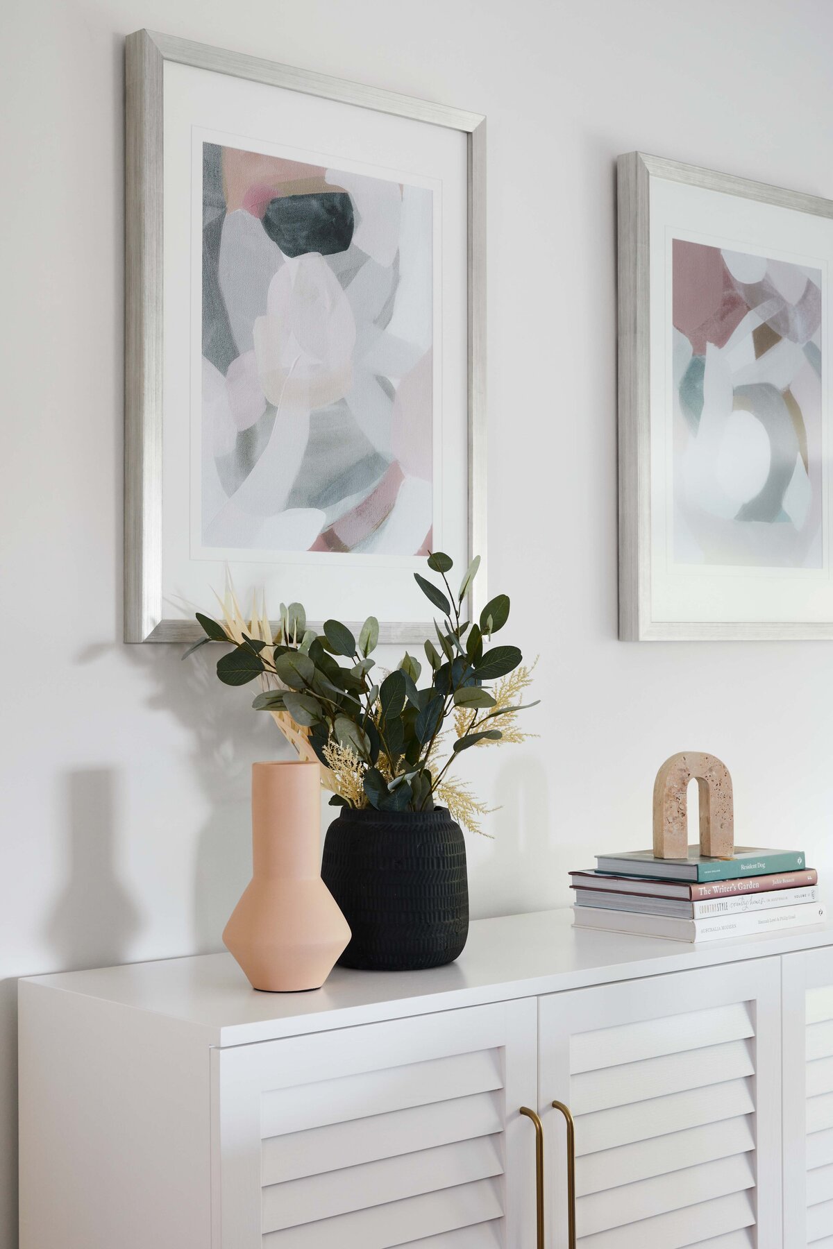 Close-up of white sideboard with natural and black ceramic vases, sculptural bookends and stacked interior books under framed abstract artwork.