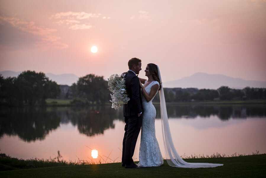 A bride and groom embrace in front of a lake at sunset.