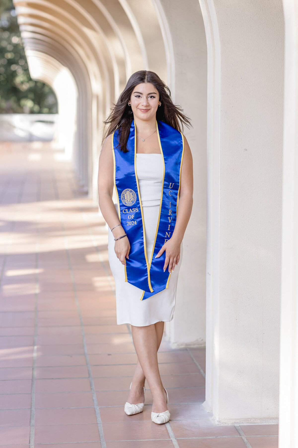 A woman in a white dress and blue graduation stole stands in a corridor with arches. She is smiling and has long, dark hair. The stole is embroidered with the year 2023 and University. The setting is bright and sunny.