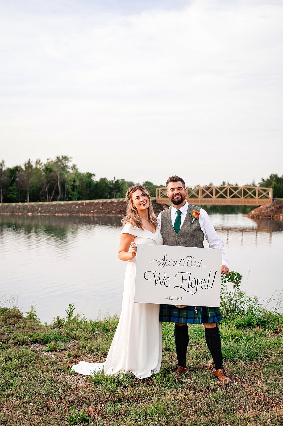 Scottish bride and groom hold up a sign that reads, "Secret's Out...We Eloped!" The groom is wearing a white shirt, grey vest, green tie and a green and blue kilt. The bride is wearing a fitted, off the shoulder wedding dress with a short train. They are standing lakeside at Sugarfoot Farm near Nashville.