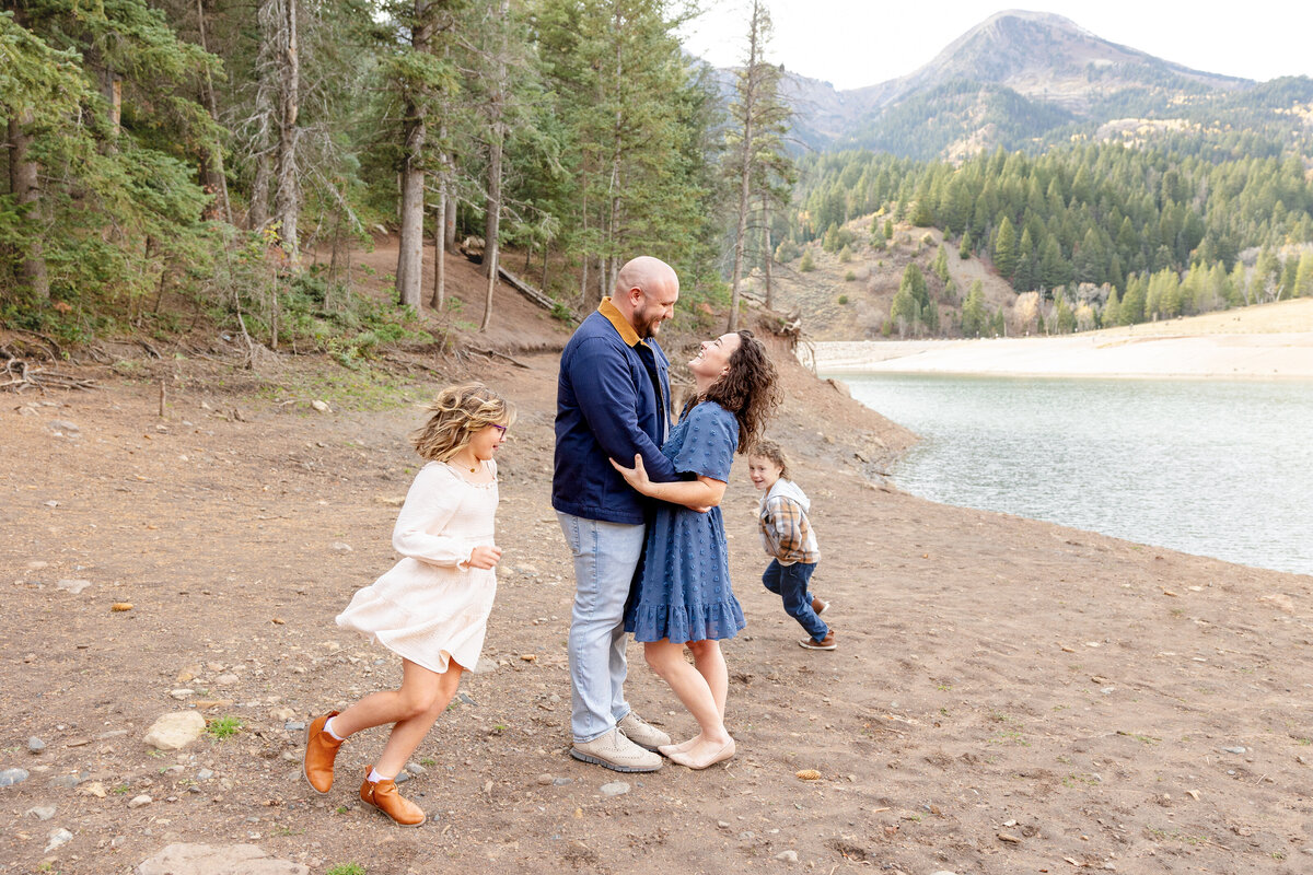 A mother and father hold each other and laugh while their two children run around them, playing near the water’s edge at Tibble Fork Reservoir in Utah.