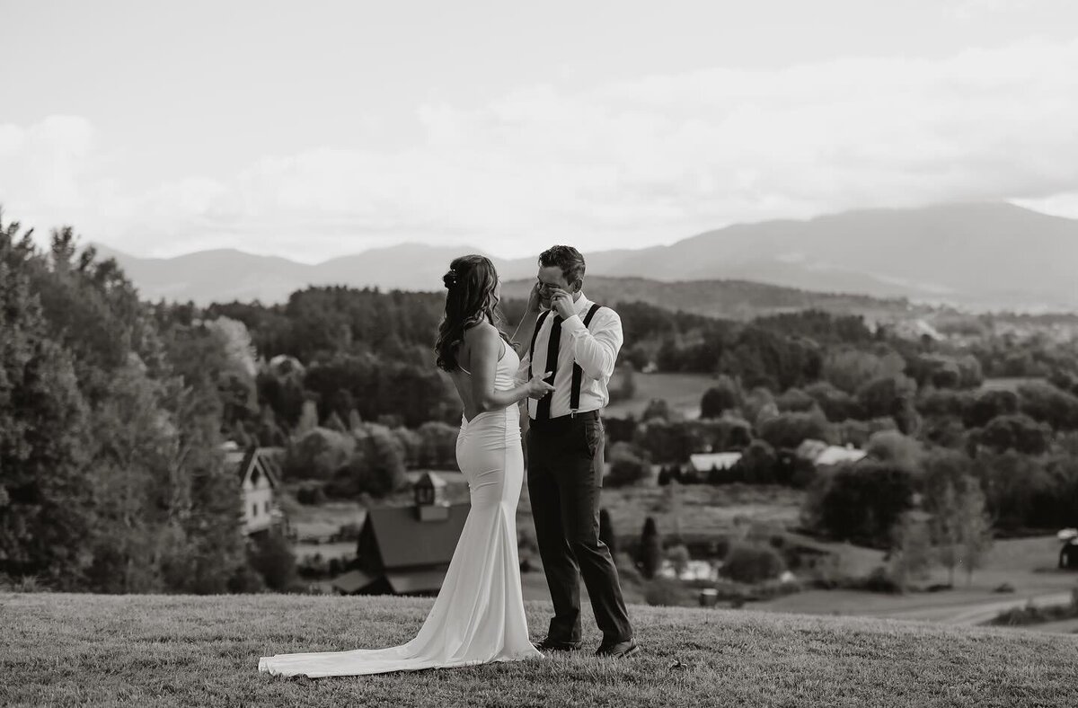 A bride in a long white dress and a groom in shirt and suspenders share an elopement moment outdoors. On a grassy hill, with New England's scenic trees, rolling hills, and cloudy skies as their backdrop, they gaze at each other. The black-and-white image captures timeless romance.