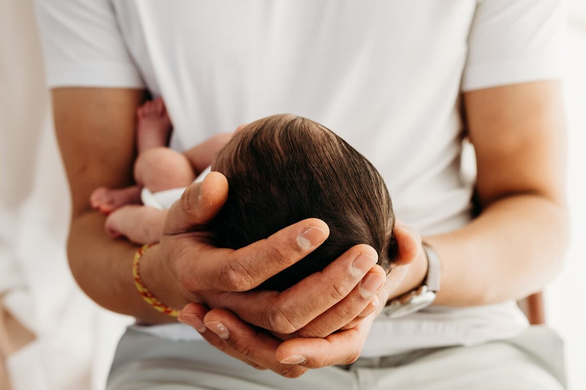 detail of newborn baby girl in her dads hands. dad wears white and baby is wrapped in a white swaddle