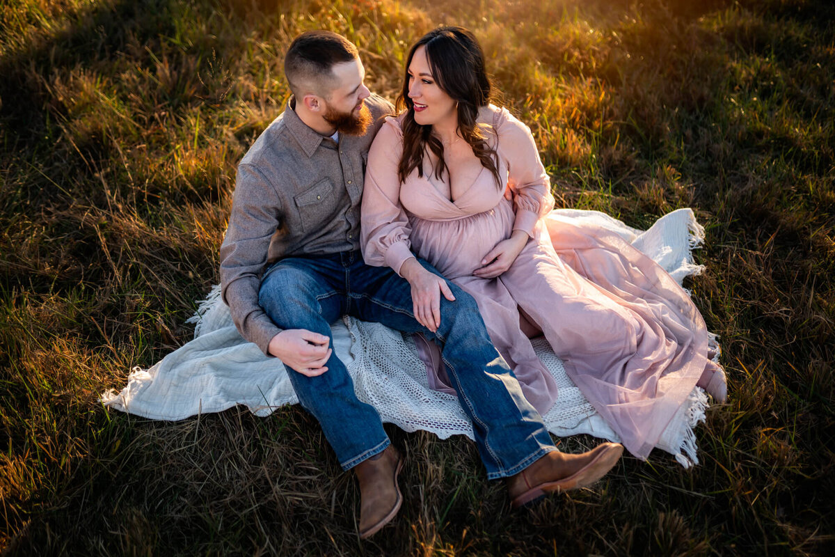 An adorable expecting couple snuggles on a blanket in the grass