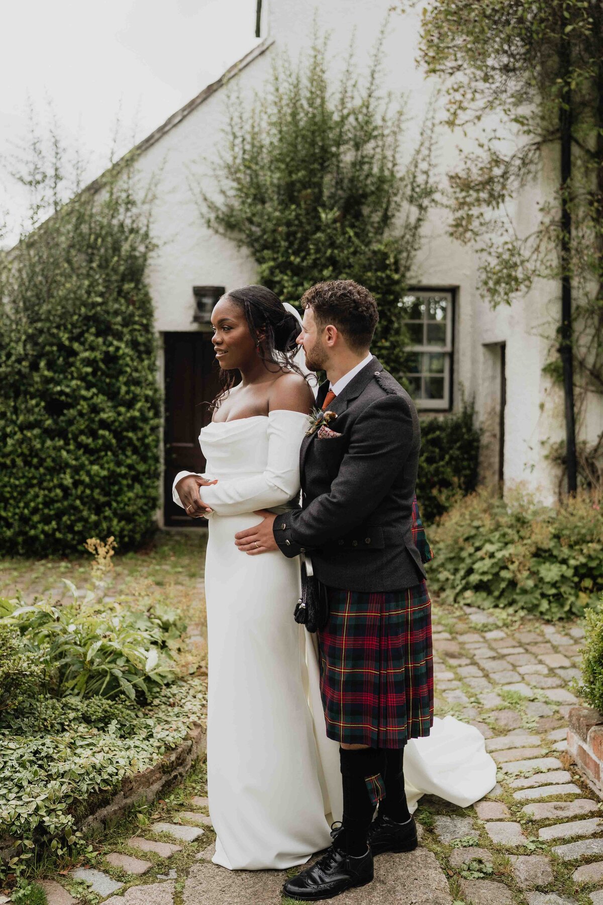 A couple stand side by side in the courtyard of Logie Country House in Aberdeen. The are photographed smiling at each other while they both wear wedding attire.