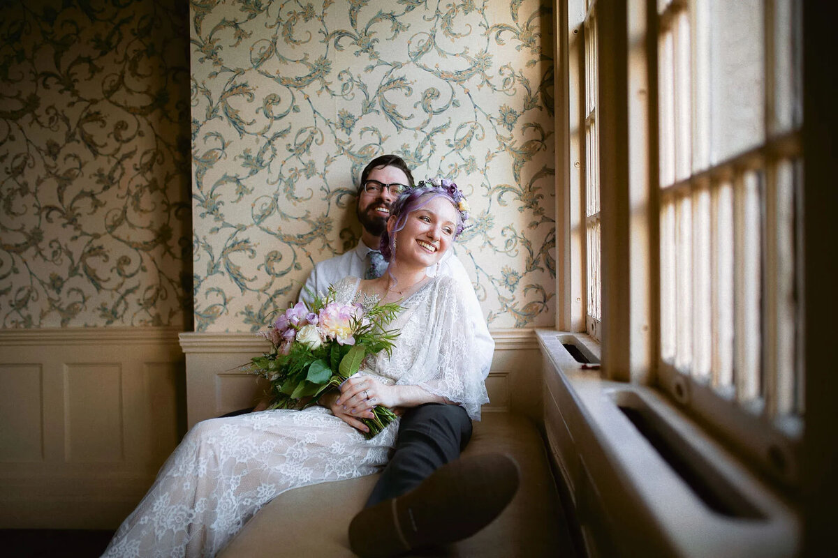 A smiling couple sits together by a window; the woman in a lace dress and flower crown holds a bouquet, while the man sits closely behind her. Captured by an NJ wedding photographer, both look happy and relaxed in a softly lit, vintage-style room.
