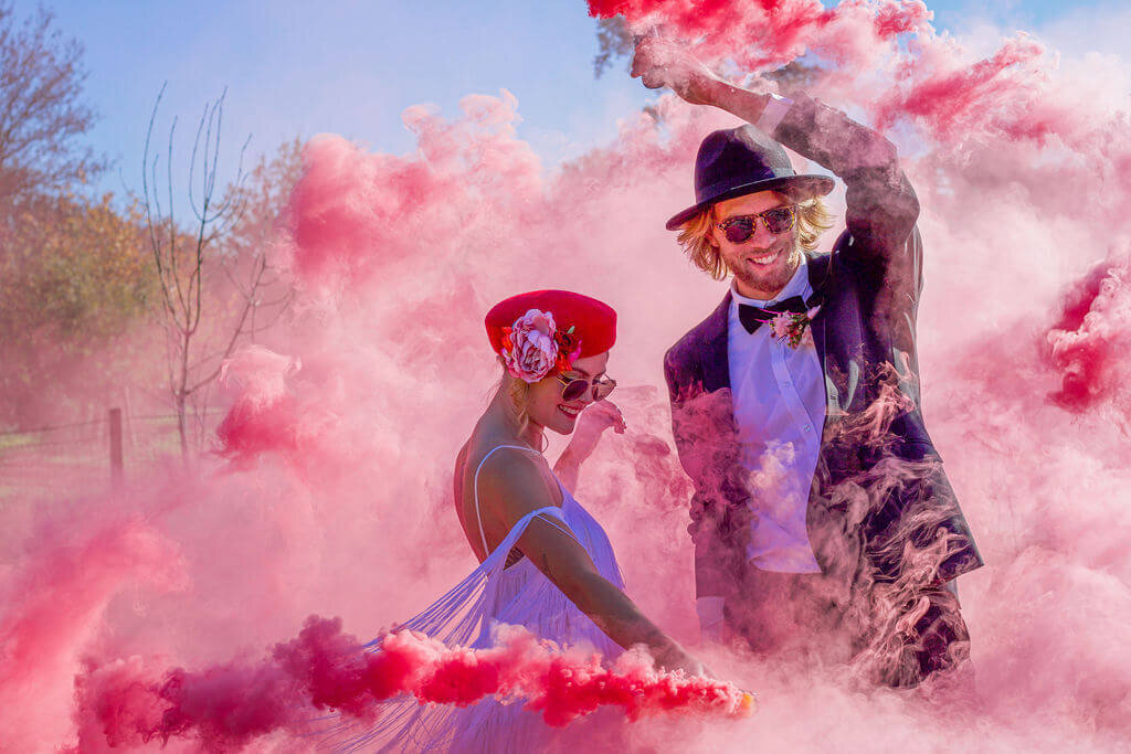 Bride in hat with groom in hat using pink smoke grenades