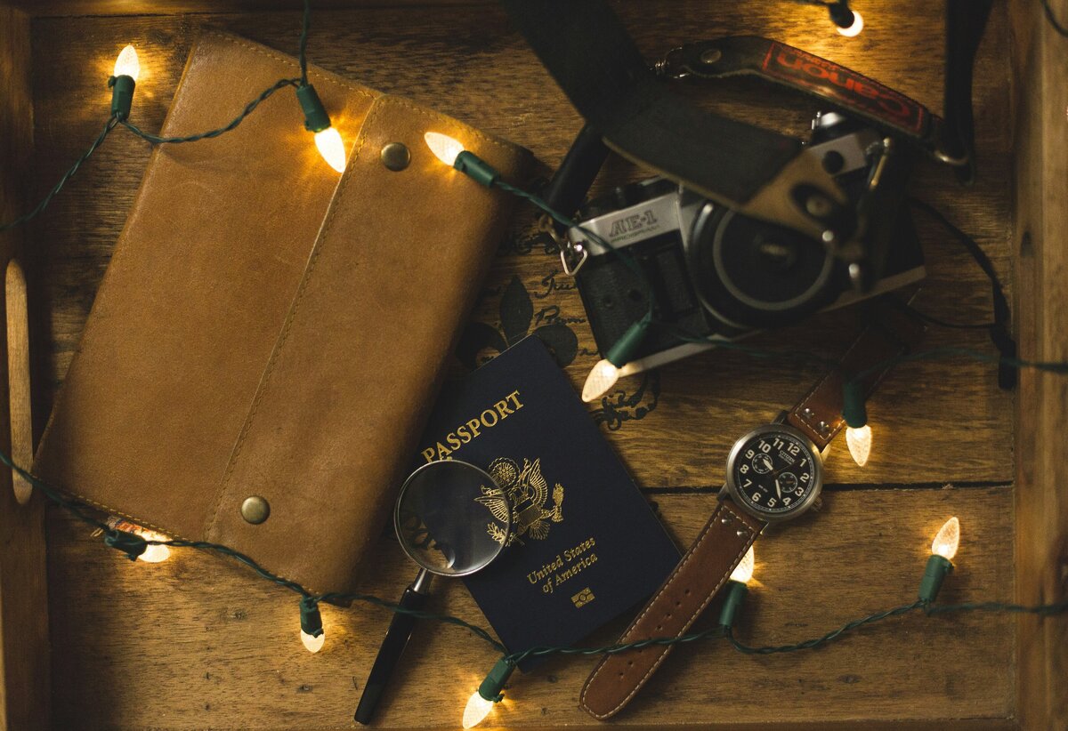 Vintage passport, journal, camera and watch illuminated by white fairy lights on rustic desk. 