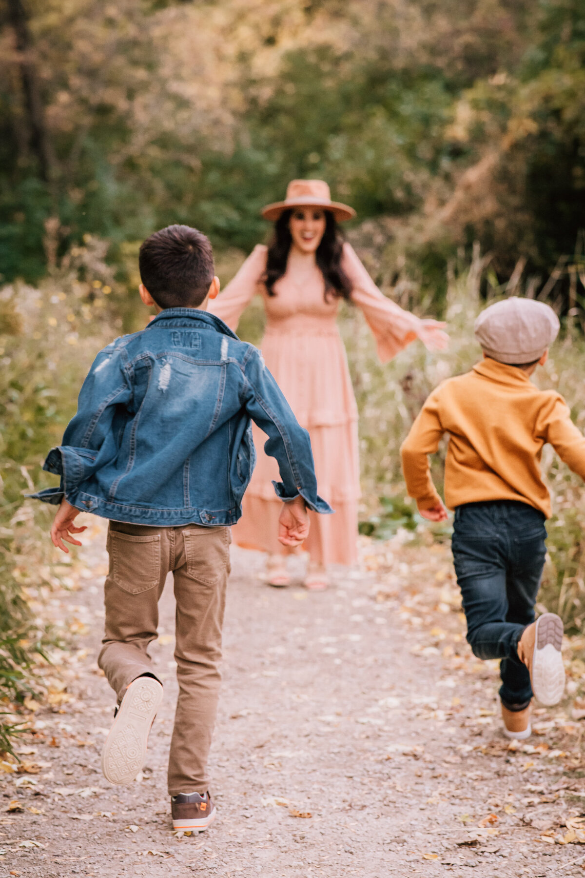 Two young boys run eagerly towards their mother, who is standing at the end of a trail with open arms, wearing a long dress and hat. The boys are seen from behind, with one in a denim jacket and the other in a yellow sweater.