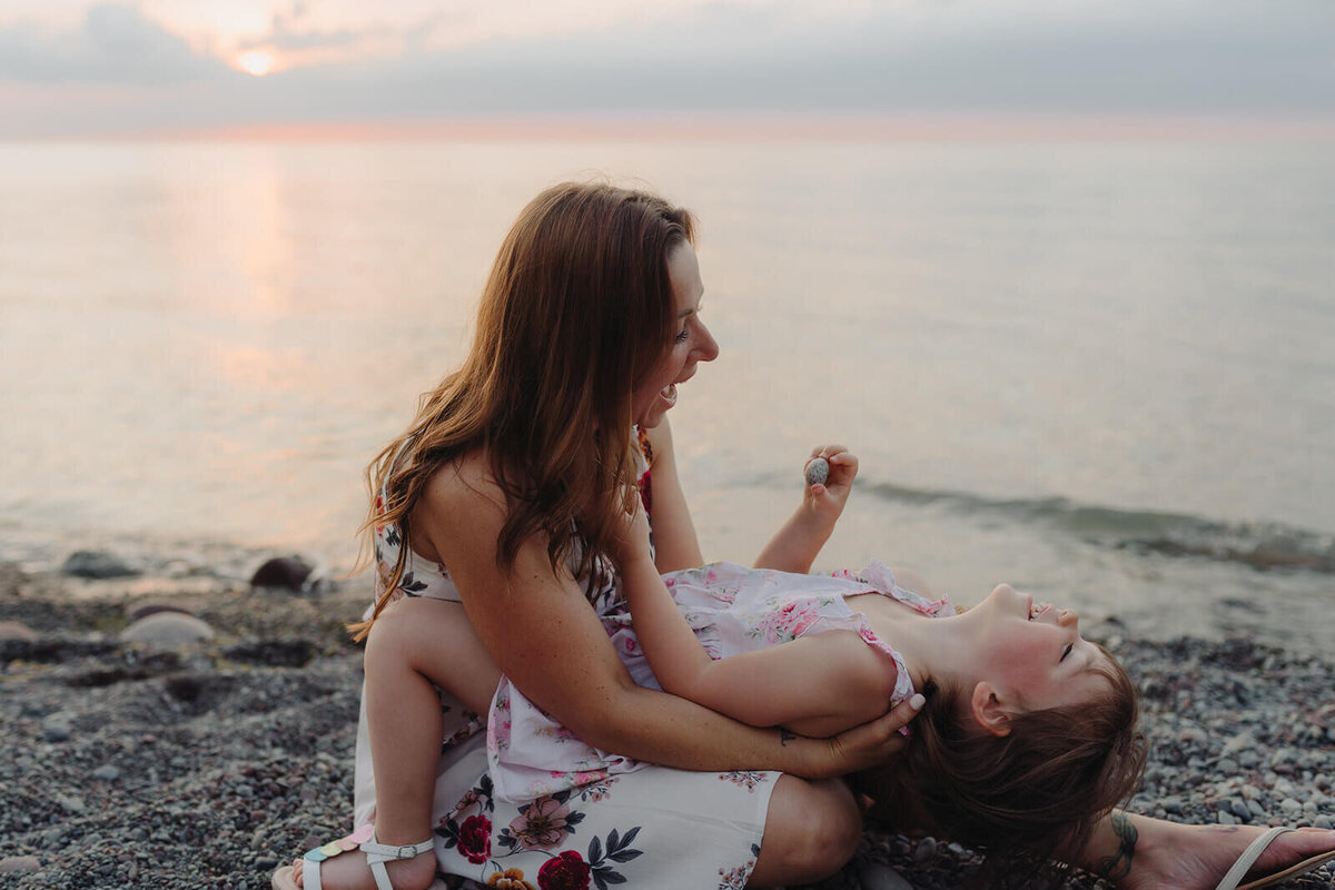 woman with laughing little girl in her lap at family with two kids on the beach at family photo shoot in Finger Lakes, NY