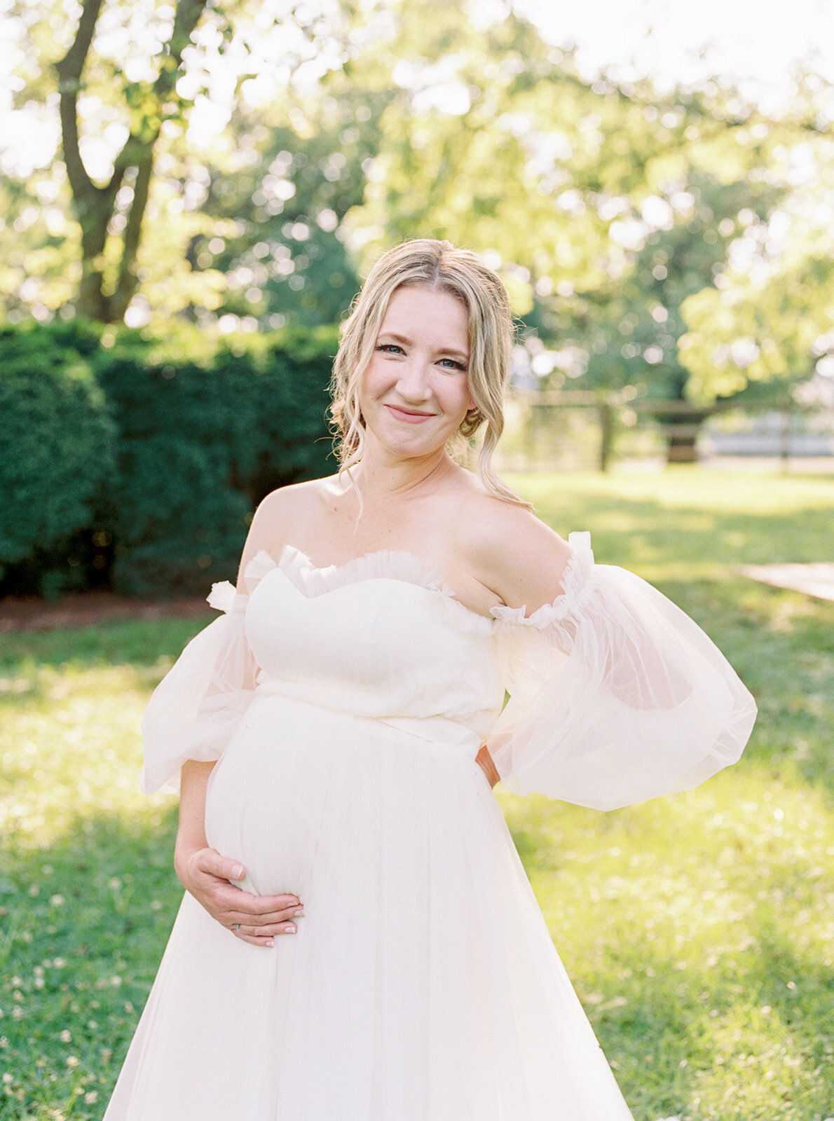 Blonde pregnant mother wears a white flowing gown and holds her belly with one hand and places her other hand behind her back while smiling at the camera