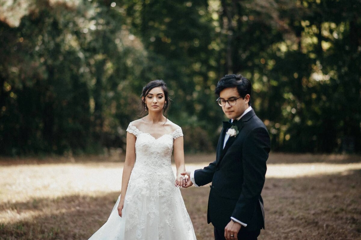 A bride and groom holding hands and looking back, with a wooded area behind them, in a poised and elegant wedding portrait.