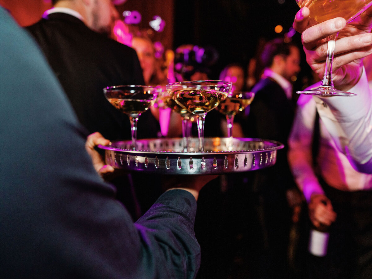 A waiter carries a tray of elegant champagne coupe glasses as guests celebrate in the background.