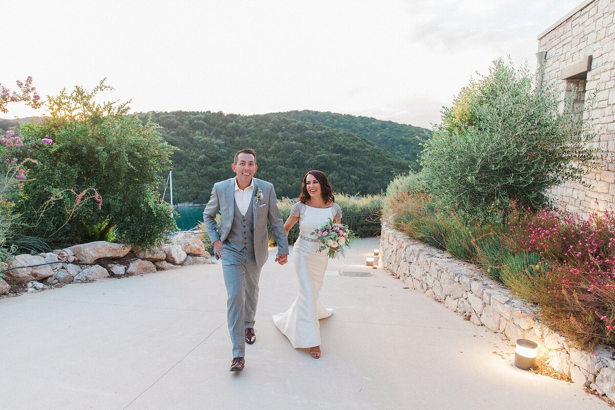 Bride and groom walk together through a scenic hilltop venue surrounded by olive trees, smiling during their relaxed destination wedding in Greece