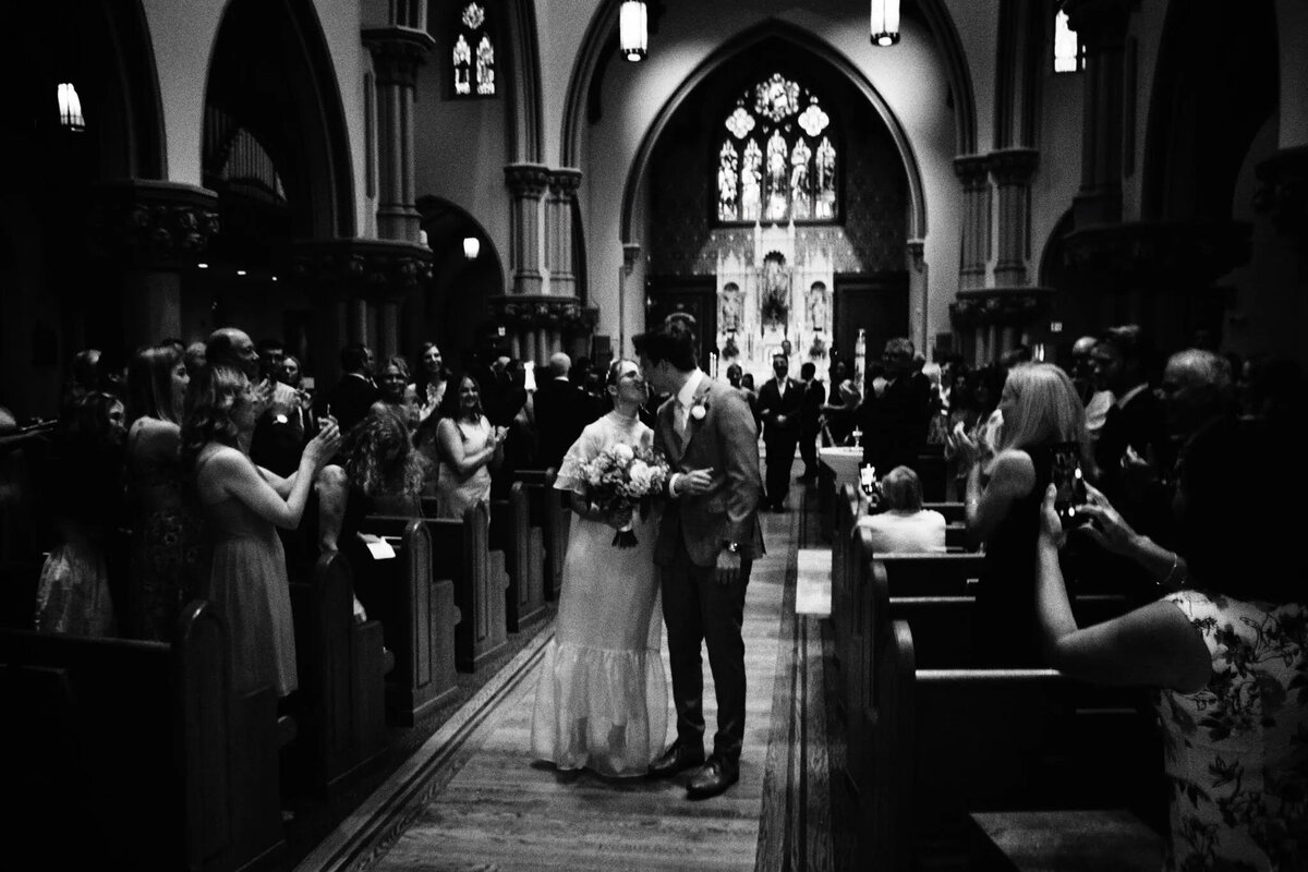 A bride and groom walk down the aisle of a gothic-style church, surrounded by guests celebrating their wedding. Captured in black and white by an NJ wedding photographer, this candid moment radiates pure joy and timeless elegance.