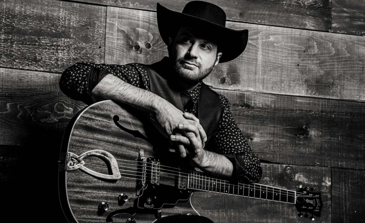 Country music photo Doc Reinhart black and white wearing cowboy hat sitting with guitar hands together wood wall background