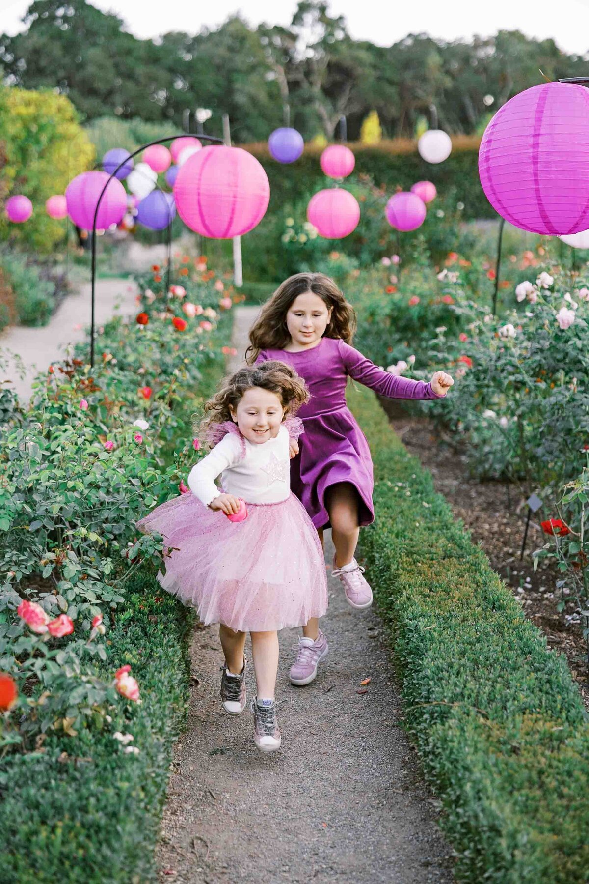 two little girls wearing pink dresses and running through a rose garden
