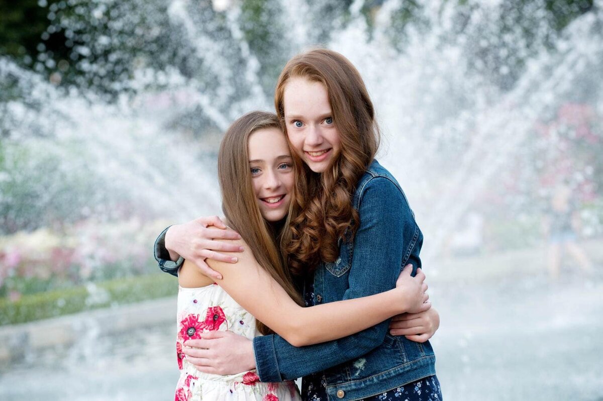 2 Sisters happily hugging each other and in front of a water fountain in the park.
