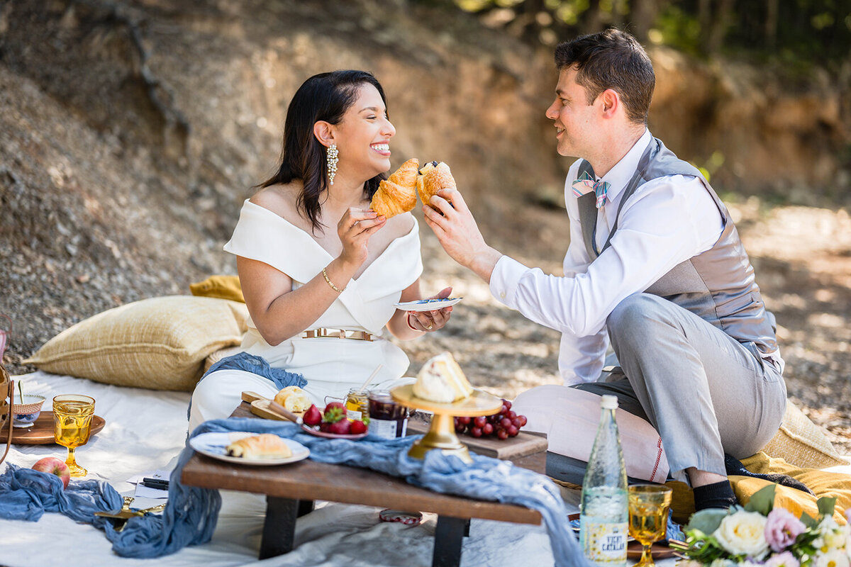 A couple sits down for a brunch picnic on their elopement day along the shores of Carvin’s Cove in Roanoke, Virginia. The picnic setup includes a picnic basket, a couple of pillows, a small table with a draping blue fabric, and all of their food, including croissants, various fruits, cakes, jam, and honey. The bride and groom “clinks” croissants and laughs.