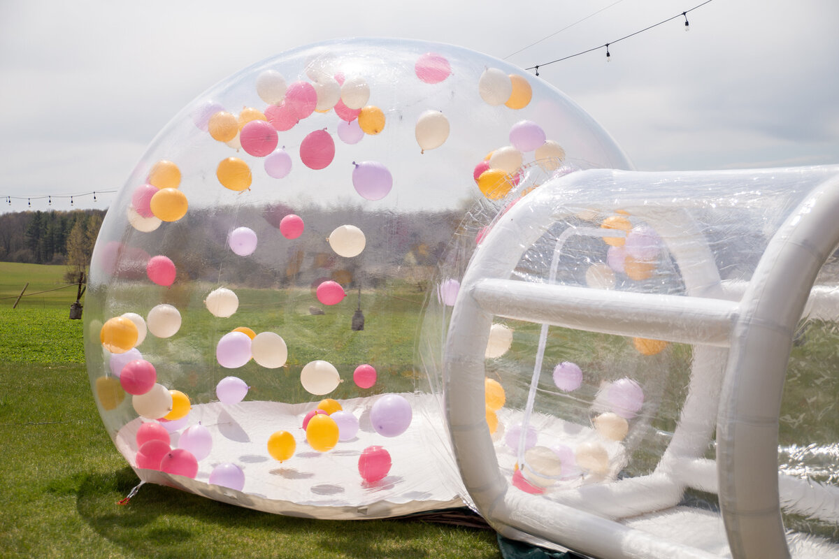 A large transparent bubble set up on the grass with colorful balloons flying around inside the dome.