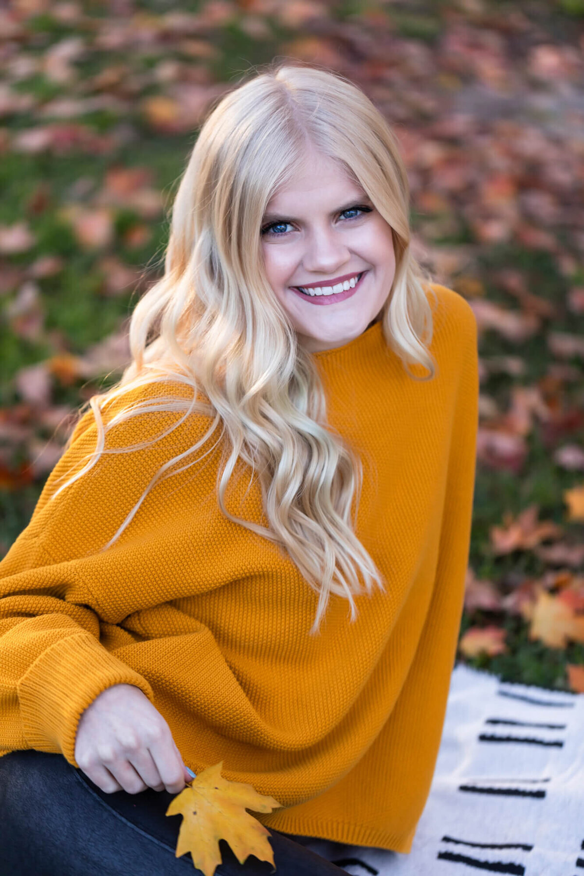 A beautiful blonde teen poses for a senior portrait wearing an orange sweater and holding a golden Fall leaf. Captured by Springfield, MO senior photographer Dynae Levingston.