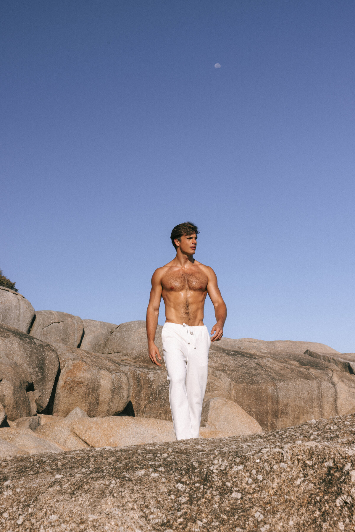 Shirtless male model walking across sunlit rocks in Cape Town, wearing white drawstring pants under a clear blue sky with the moon faintly visible above.