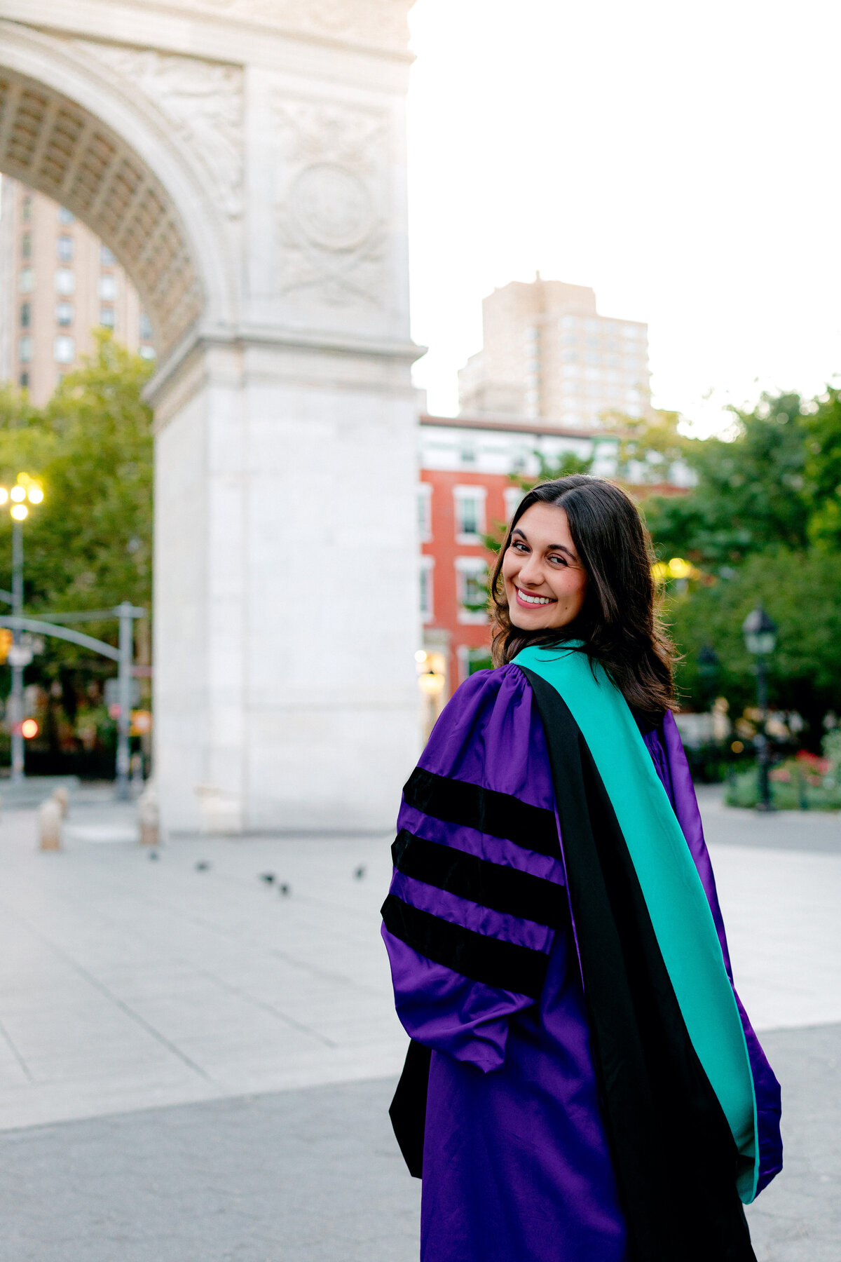 NYU graduate smiling over her shoulder in purple gown and hood at Washington Square Park during sunrise.
