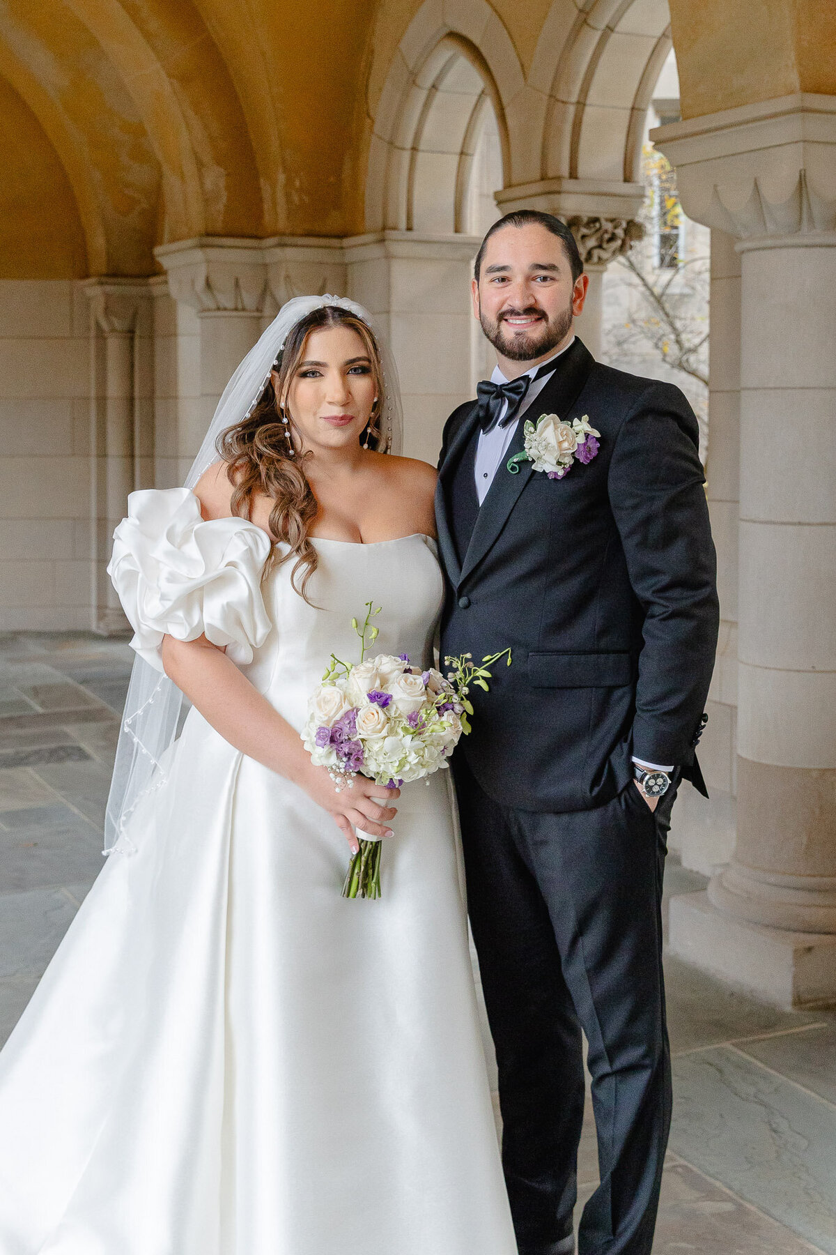 A bride and groom pose for a portrait. Behind them is lovely architecture. They both are smiling at the camera.