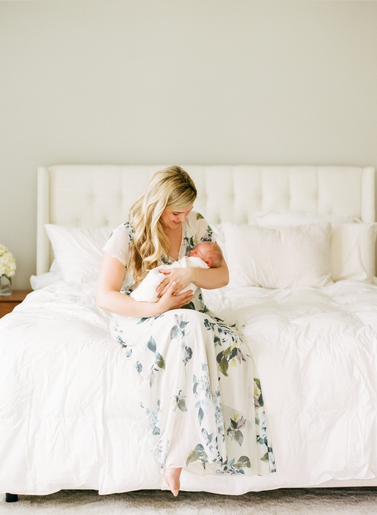 Mom sits on the end of her bed and holds her newborn during a Holly Springs Newborn session. Photographed by Raleigh Newborn Photographer A.J. Dunlap Photography.