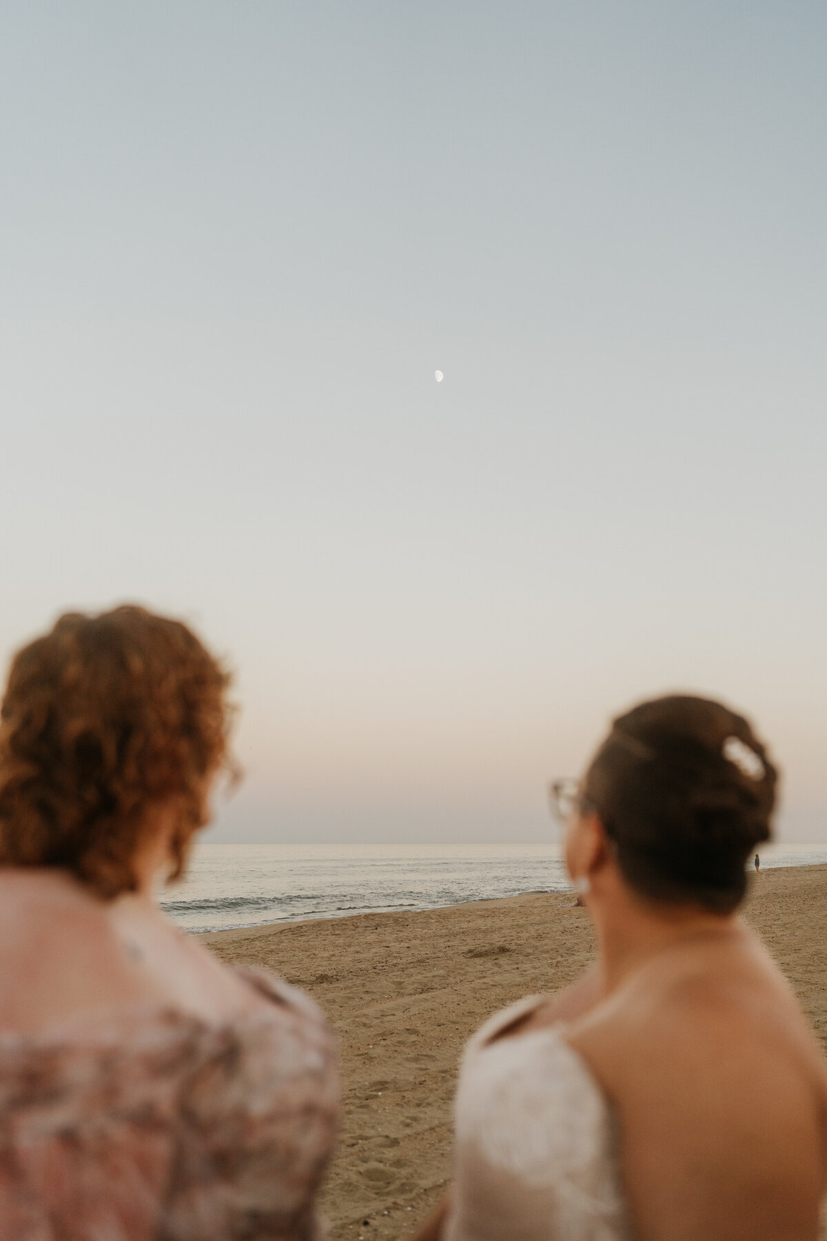 A newlywed couple standing close and looking off into the distance at the beach 