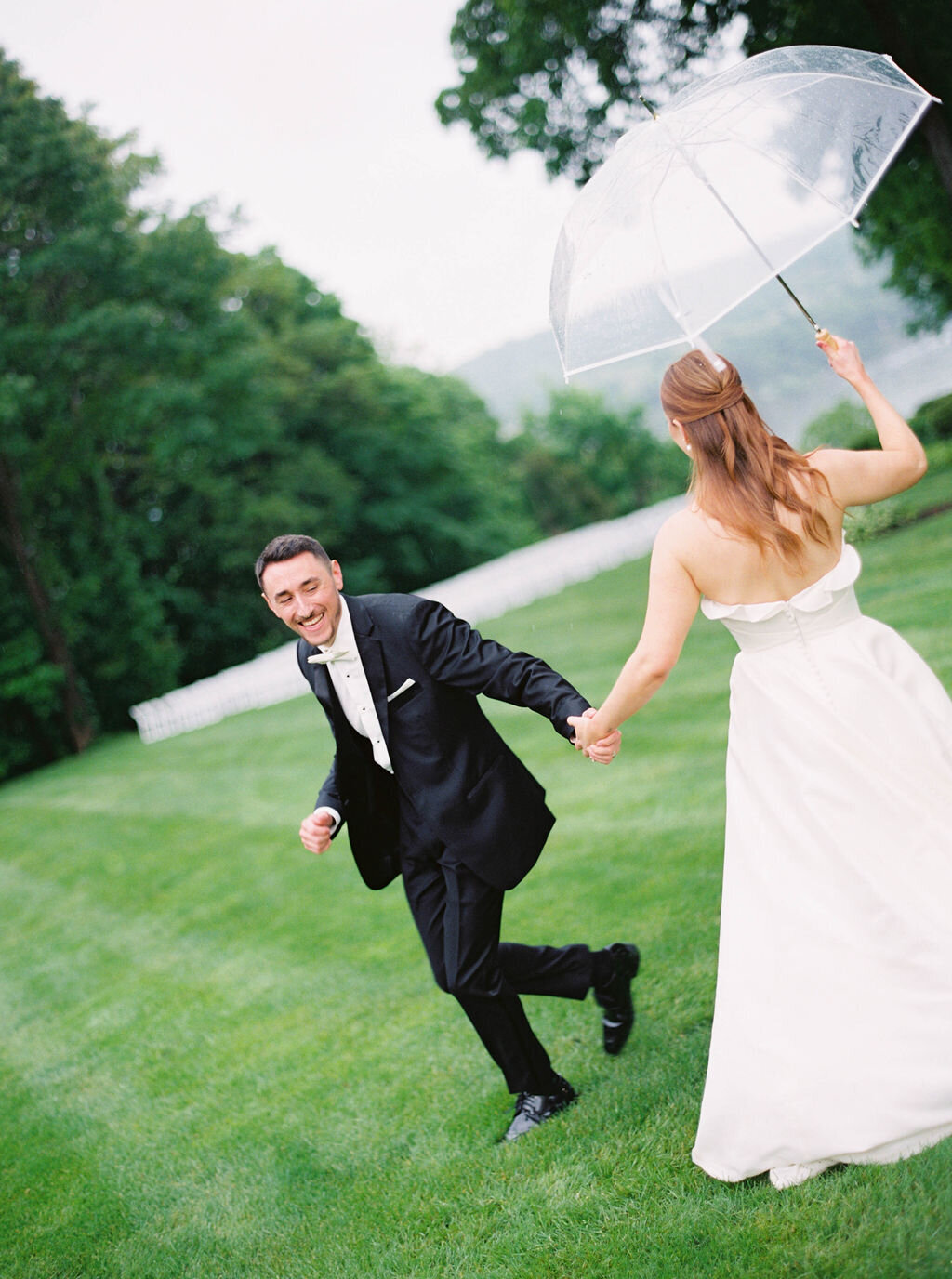 Wedding Photographer RI | A bride and groom joyfully run hand in hand on lush green grass. The groom wears a black suit, while the bride holds a clear umbrella, her dress flowing.