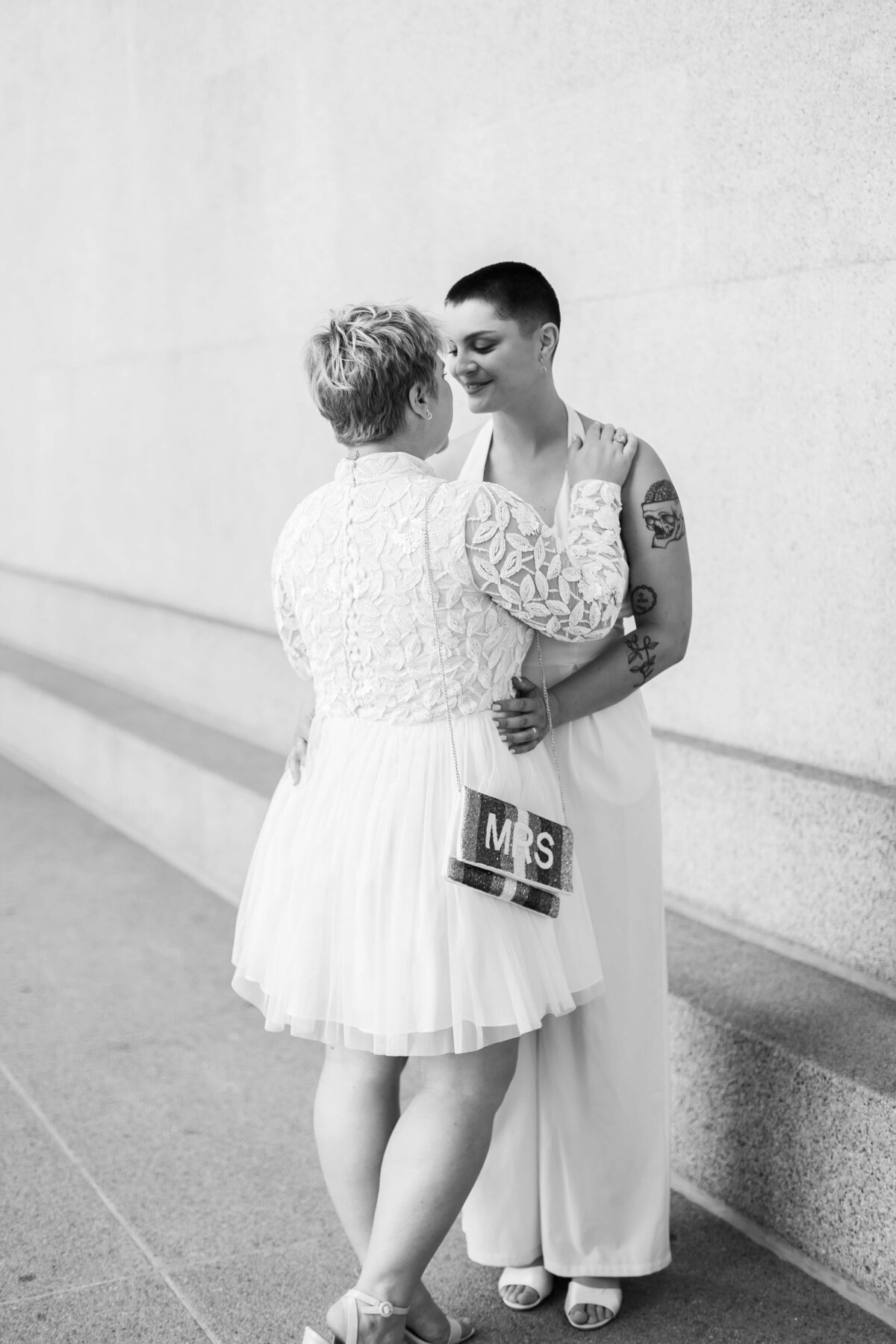 One bride wears a white jumpsuit and the other wears a long sleeve lace dress holding a Mrs bag