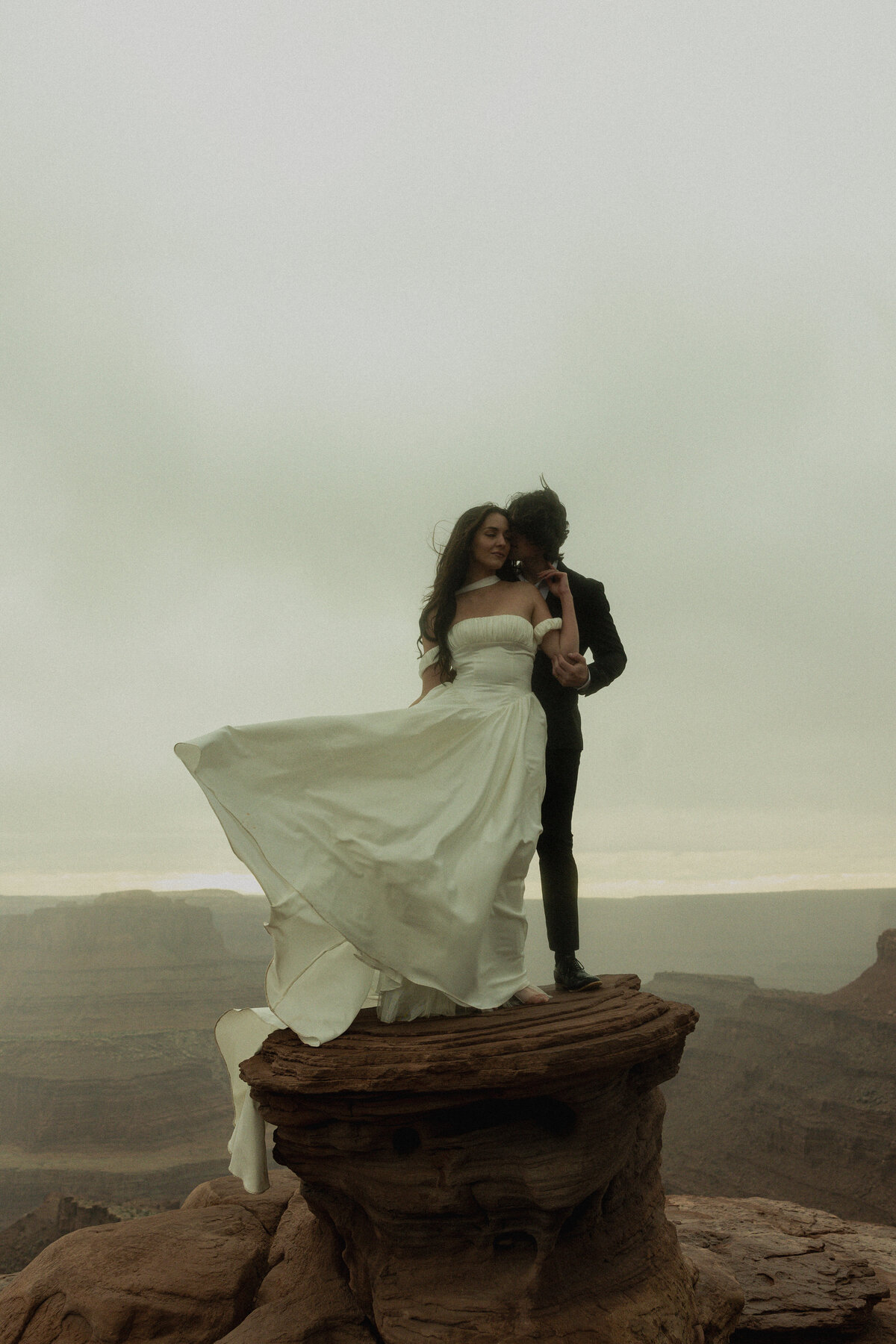 A couple in wedding attire embraces on a rocky outcrop, with dramatic clouds and stunning canyon vistas in the background.
