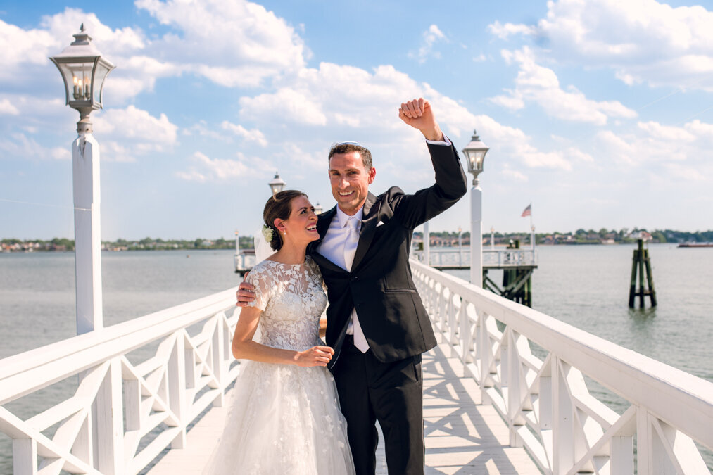A bride and groom standing on a pier with their arms raised.