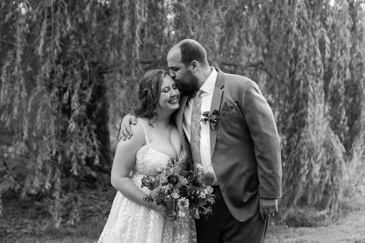 Bride-and-groom-kiss-in-front-of-willow-trees-at-forest-outdoor-venue-Twin-Willow-Gardens-in-Snohomish-WA-photo-by-Joanna-Monger-Photography